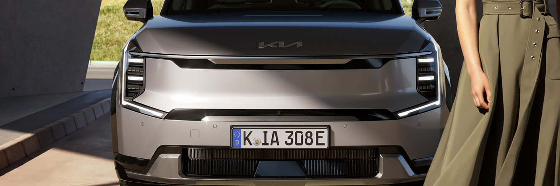 Kia EV9 von vorne; am Bildrand ist eine Frau in einem dunkelgrünen Kleid angeschnitten zu sehen.
