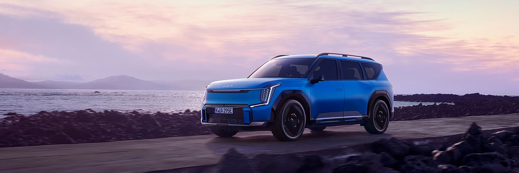 Kia EV9 GT-Line fährt auf einer Landstraße am Meer im Sonnenuntergang.